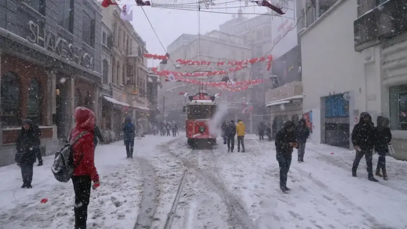 Yoğun kar yağışı altında İstanbul’un İstiklal Caddesi; karla kaplı raylarda ilerleyen kırmızı nostaljik tramvay ve karda yürüyen insanlar