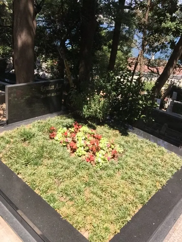 Kanlıca Mezarlığı’nda Kayahan Açar’ın çimle kaplı, ortası çiçekli mezarı; siyah granit taşta adı ve doğum ile ölüm tarihleri, arkada ağaçlar arasından görünen Boğaz