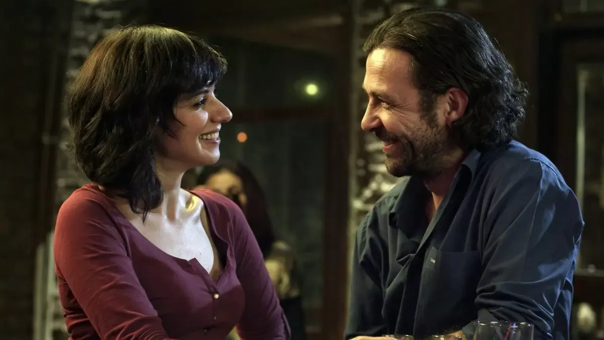 Kaybedenler Kulübü filminden, bir barda birbirine gülümseyerek bakan Zeynep ve Kaan