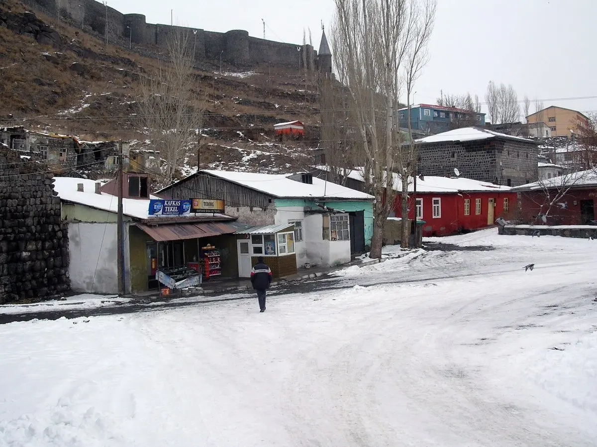Kars’ta karla kaplı bir sokak; kar örtülü çatılı tek katlı dükkânlar ve kırmızı boyalı eski taş evler, arkadaki tepede Kars Kalesi’nin surları ve karlı yolda yürüyen bir adam