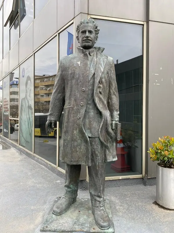 İstanbul Şişli’de cam cepheli bir binanın önünde, paltosu açık ve elleri ceplerinde duran bronz Nâzım Hikmet heykeli