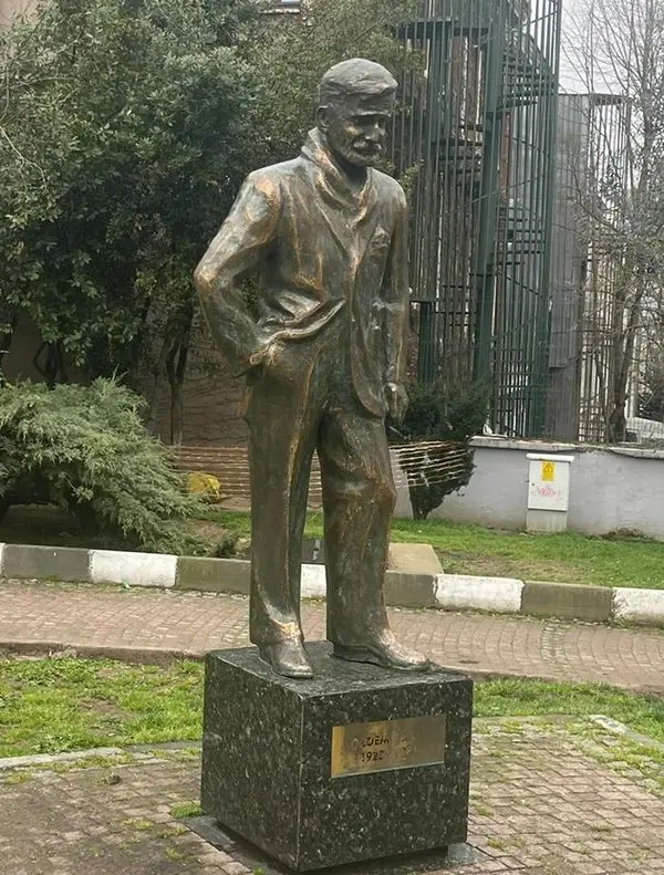 Ağaçlıklı bir alanda kaide üzerinde duran, ceketli ve şapkalı Özdemir Asaf heykeli