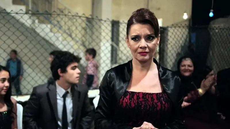 Köksüz (2013) filminden bir kare: tel örgülü bir avluda kurulan kalabalık bir eğlencede kırmızı-siyah elbiseli bir kadın ayakta duruyor, arkasında takım elbiseli genç bir oğlan ve masalarda oturan konuklar