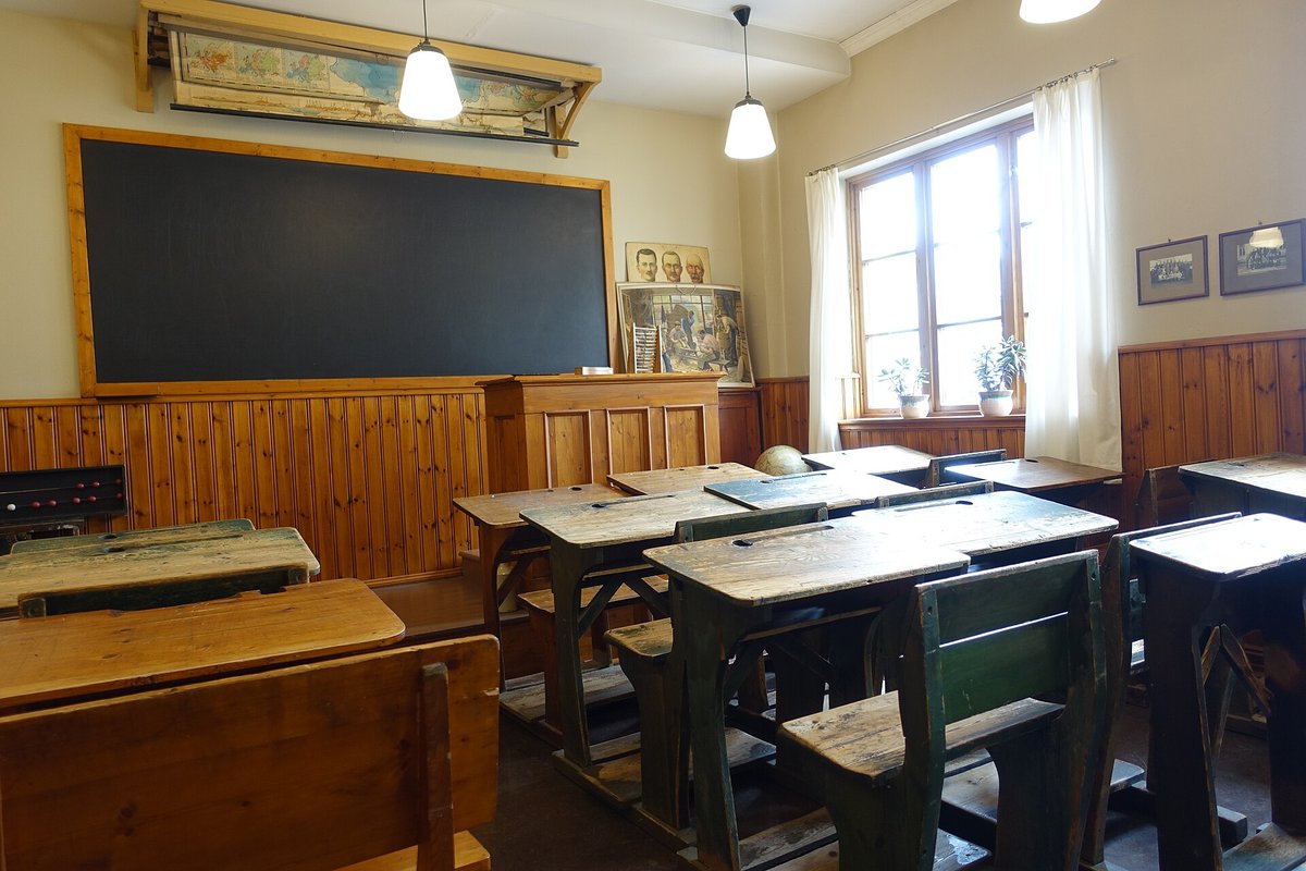 Eski bir sınıfta ahşap sıralar, büyük kara tahta, tavandan sarkan lambalar ve perdeli pencere