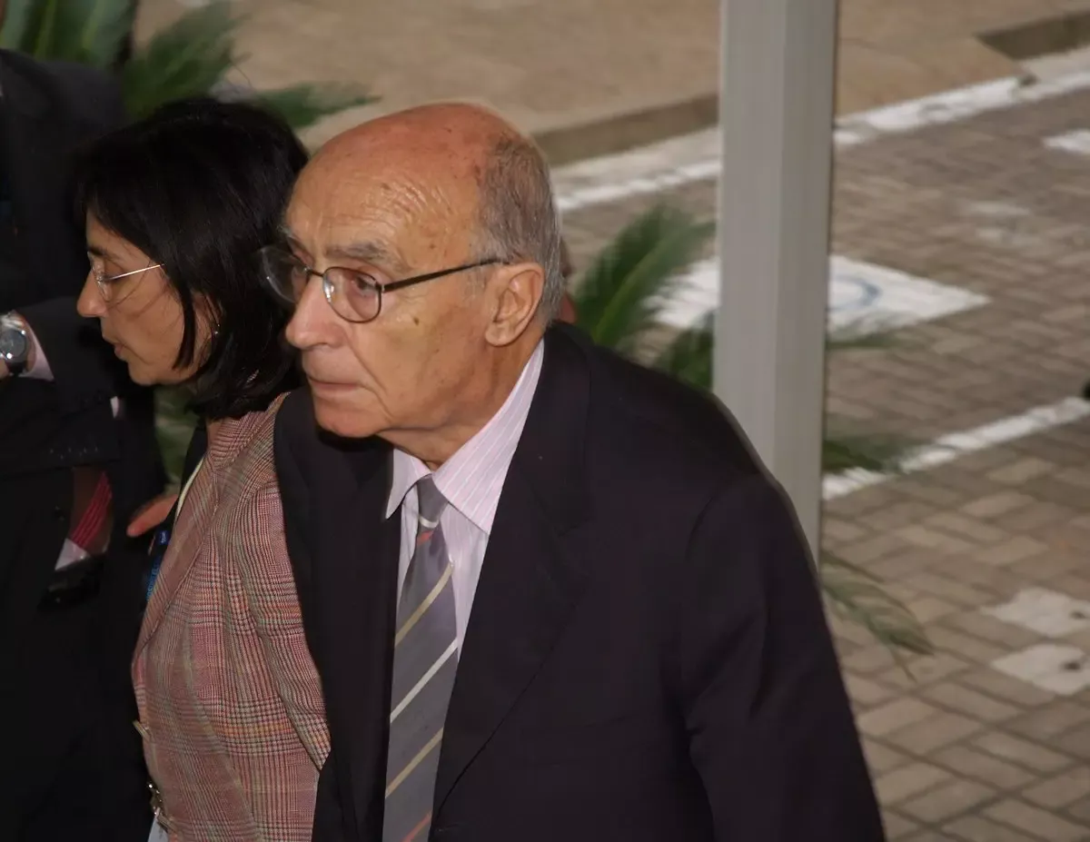 José Saramago 2005 yılında koyu renk takım elbise ve gözlüğüyle, yanında bir kadınla birlikte açık havada yürürken