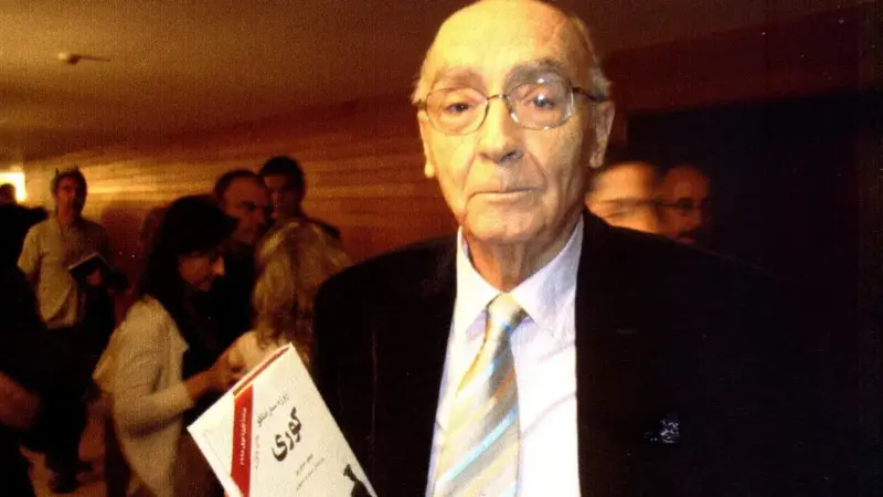 José Saramago, elinde Körlük romanının Farsça çevirisiyle, kalabalık bir salonda objektife bakarken