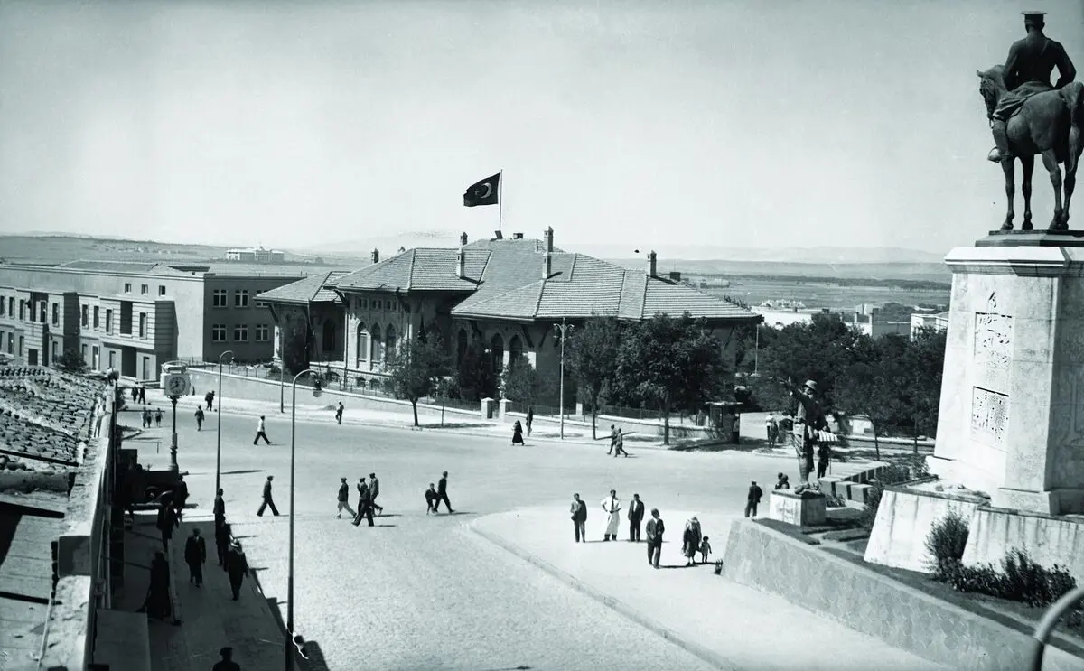 Ankara Ulus Meydanı'nda sağda kaidesi üzerinde atlı Zafer Anıtı, karşıda bayraklı Birinci Meclis binası ve meydanda yürüyen insanlar, siyah-beyaz fotoğraf