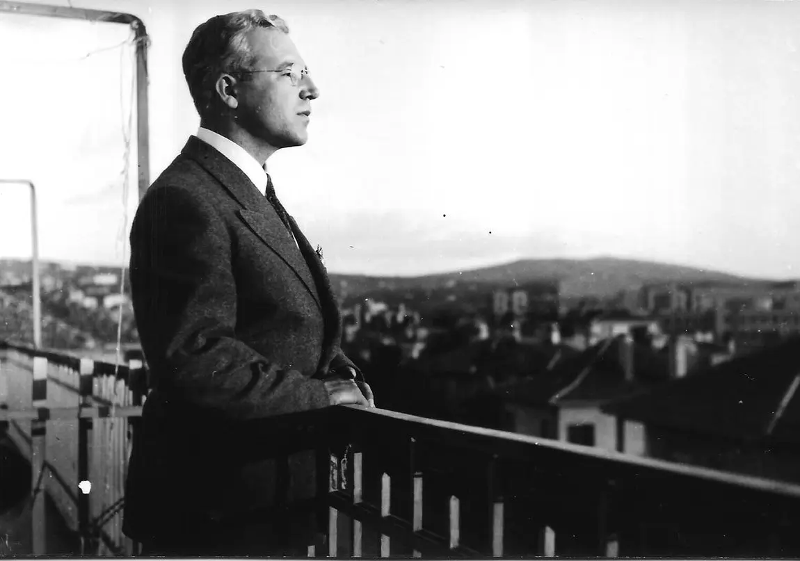 Sabahattin Ali takım elbise ve gözlükle bir balkonda durmuş, arkasında şehrin çatıları ve tepeler görünen siyah-beyaz fotoğraf