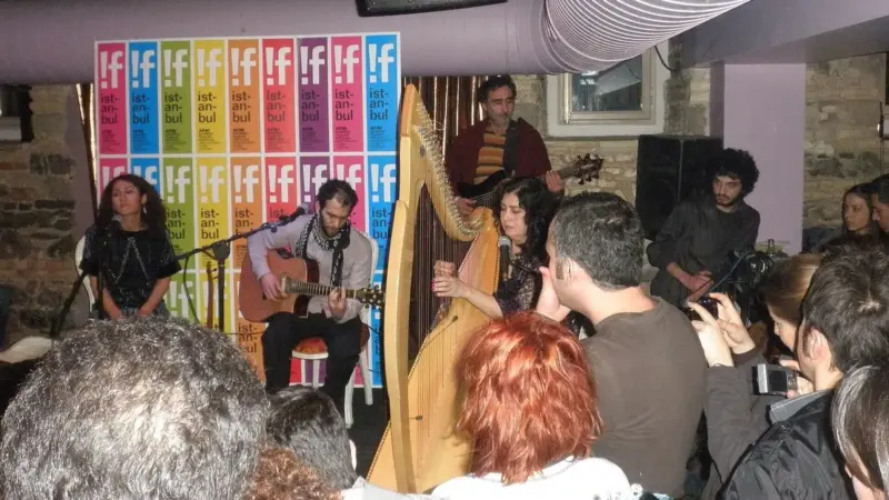 İstanbul’daki bir festival konserinde sahnede Kürtçe şarkılar seslendiren Aynur Doğan, gitarıyla Mehmet Atlı ve arp çalan Tara Jaff; önde onları dinleyen ve fotoğraf çeken seyirciler