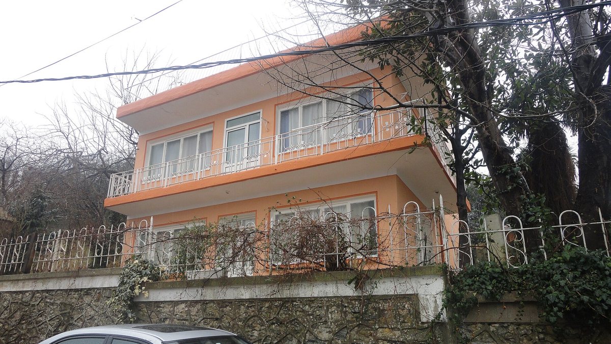 Ağaçların arasında, taş istinat duvarının üzerinde duran turuncu boyalı iki katlı balkonlu ev