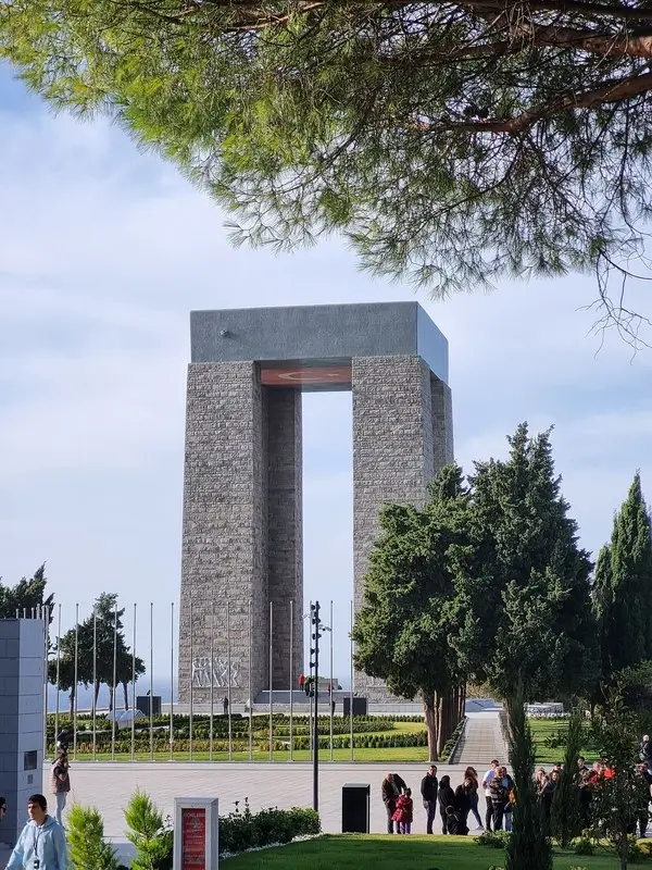 Gelibolu Yarımadası’nda çam dalları arasından görünen dört ayaklı Çanakkale Şehitleri Anıtı ve önündeki ziyaretçiler