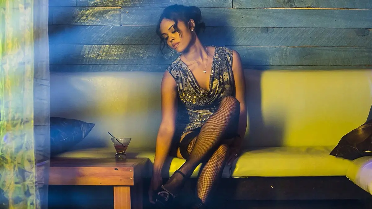 Müptela (Addicted, 2014) filminden bir kare: Zoe rolündeki Sharon Leal, desenli kısa bir elbise ve file çoraplarla loş, mavi ışıklı bir mekânda sarı bir kanepenin kenarına oturmuş, başını yana eğmiş; yanındaki ahşap sehpada bir kokteyl kadehi duruyor
