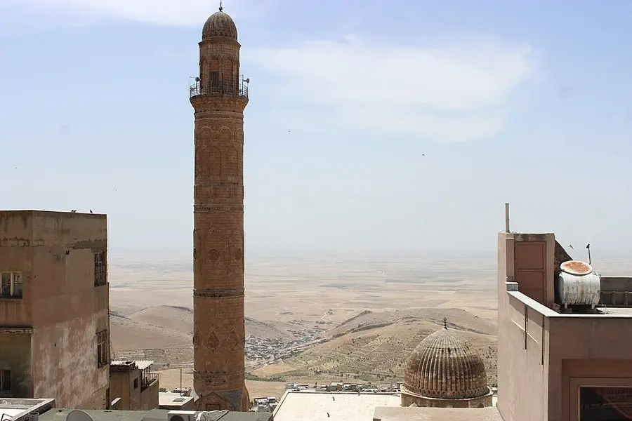 Mardin'de taş evlerin arasında yükselen eski bir minare, sağda kubbe ve arkada uzanan geniş Mezopotamya ovası