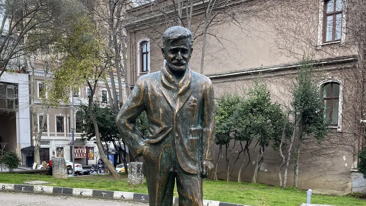 Beşiktaş Şairler Sofası’nda, ellerinden biri cebinde ayakta duran bronz Özdemir Asaf heykeli; arkada çıplak ağaçlar ve eski taş bina