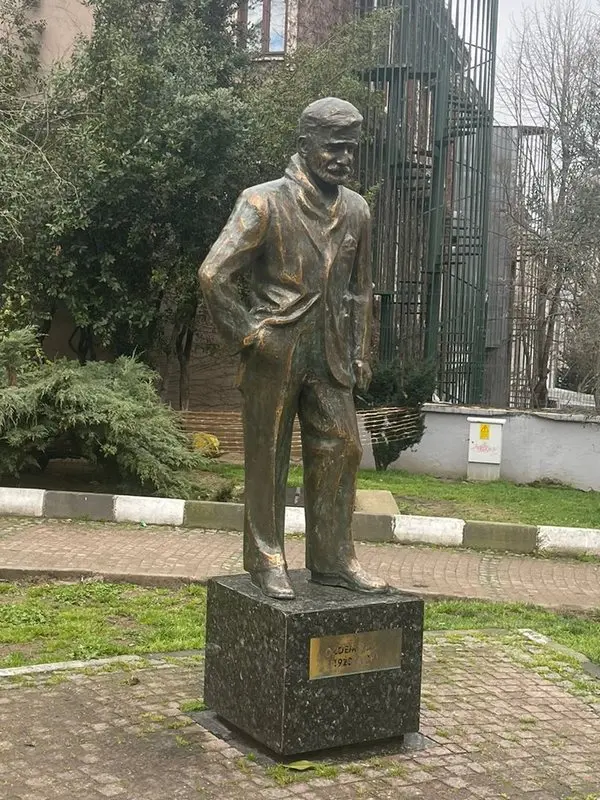 Bir parkta, taş kaide üzerinde duran, şapkalı ve atkılı Özdemir Asaf'ın bronz heykeli