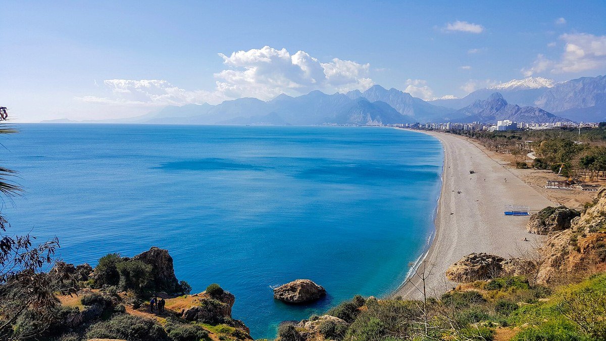 Antalya'da falezlerin üstünden görünen turkuaz deniz, uzun Konyaaltı Plajı ve arkada karlı Beydağları