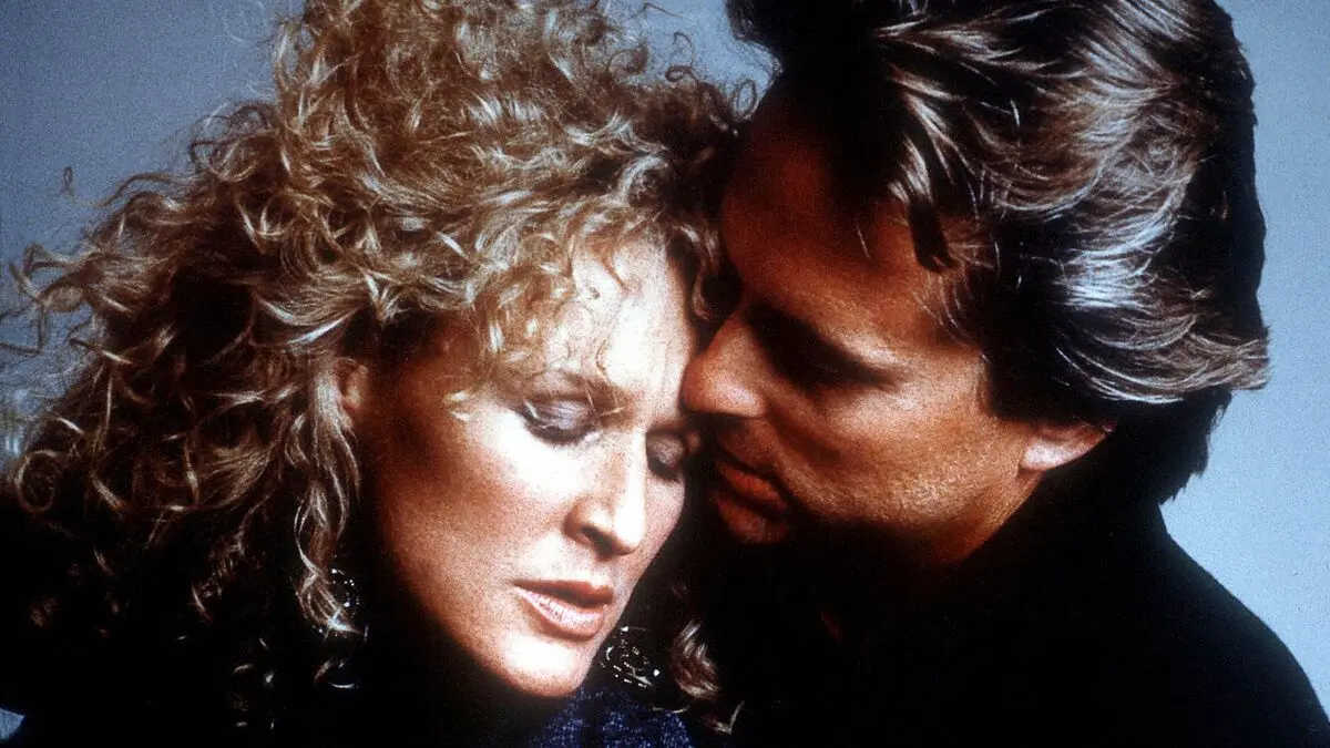 Öldüren Cazibe (Fatal Attraction) filminin tanıtım görselinde mavi gökyüzü önünde birbirine sarılan kıvırcık sarı saçlı Alex Forrest rolündeki Glenn Close ve Dan Gallagher rolündeki Michael Douglas