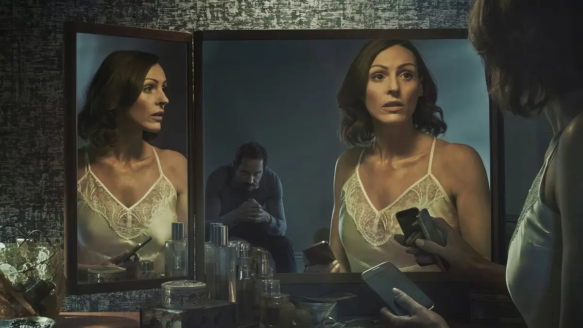 Doctor Foster dizisinden, aynalı makyaj masasının başında elinde telefonuyla duran Gemma Foster rolündeki Suranne Jones ve aynada arkada oturan Simon'ın yansıması