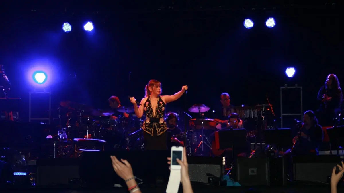 Sezen Aksu'nun İstanbul Teknik Üniversitesi konserinde mavi ışıklar altında orkestrasının önünde kollarını açarak şarkı söylemesi; ön planda telefonla çeken seyirciler