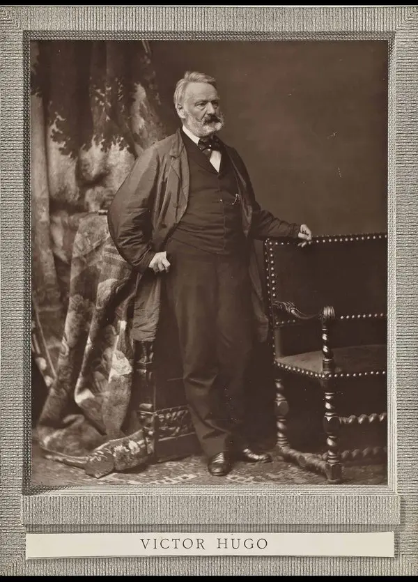 Victor Hugo’nun Bertall tarafından 1867’de çekilmiş boy fotoğrafı; koyu redingot giymiş, beyaz sakallı yazar, oymalı ahşap bir sandalyenin yanında ayakta duruyor, altta VICTOR HUGO yazısı