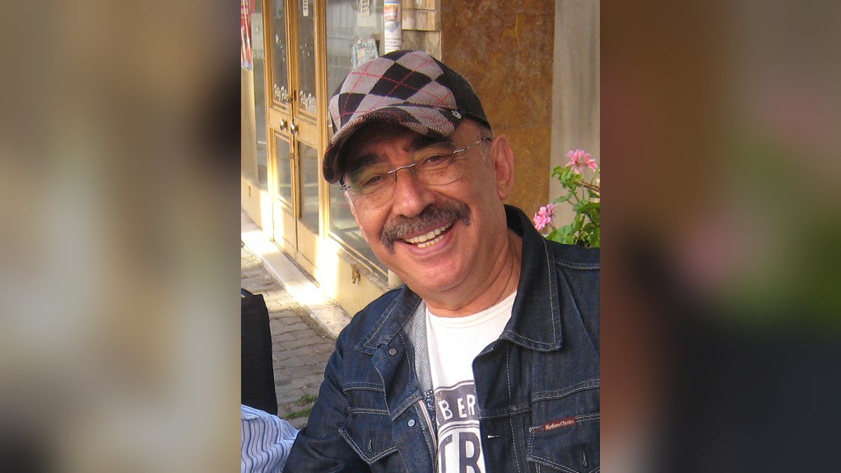 Kareli kasket ve gözlük takan, kot ceketli Şener Şen'in bir kafenin önünde gülümserken çekilmiş fotoğrafı