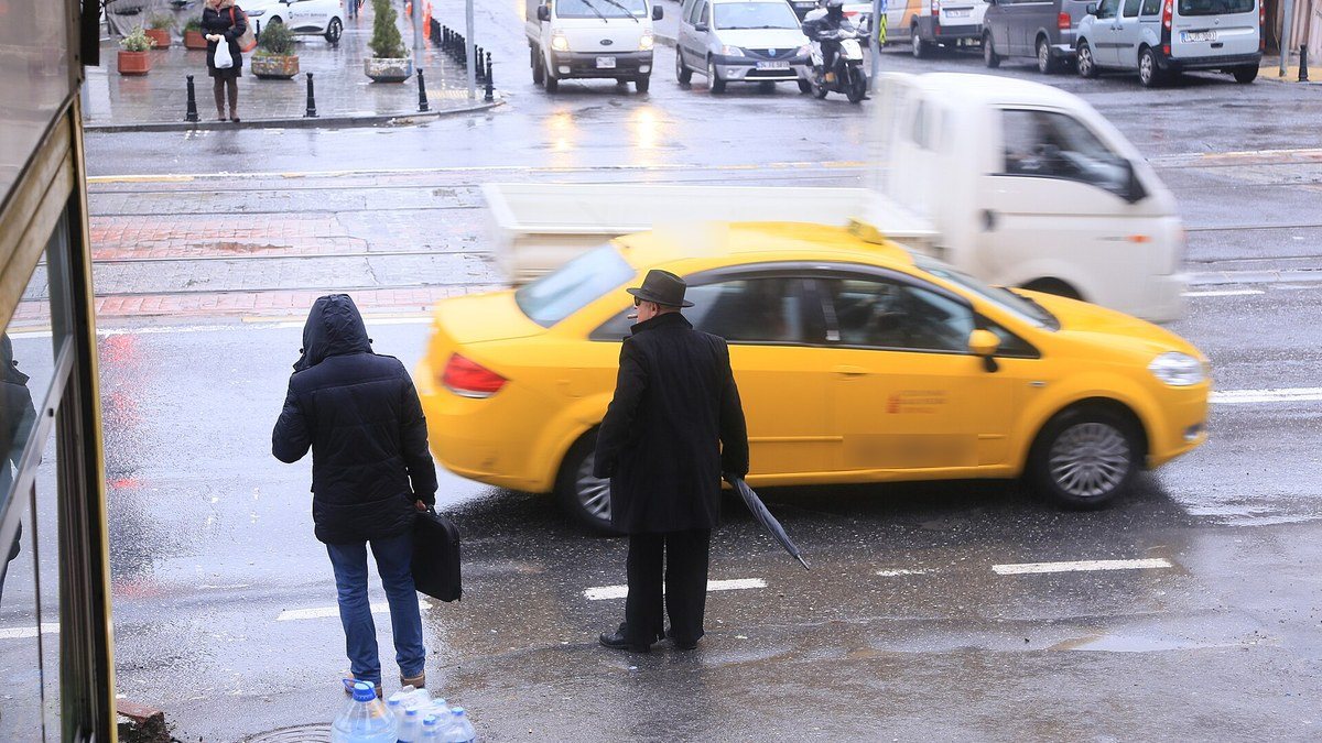Yağmurlu bir günde Karaköy'de kaldırım kenarında bekleyen fötr şapkalı yaşlı bir adam ve önünden geçen sarı taksi