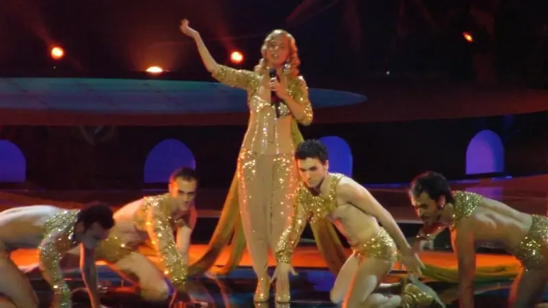 Sertab Erener, 2004’te İstanbul’daki Eurovision açılış gösterisinde altın rengi pullu kostümüyle elini kaldırmış şarkı söylerken; önünde yere çökmüş altın kostümlü dansçılar