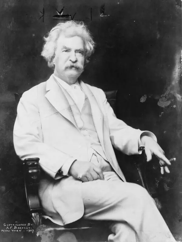 Beyaz takım elbiseli, beyaz gür saçlı ve bıyıklı Mark Twain'in bir sandalyede oturup sol elinde purosunu tuttuğu siyah-beyaz fotoğrafı
