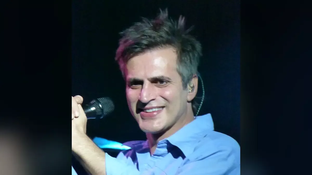 Teoman'ın açık mavi gömlekle, elinde mikrofonla sahnede gülümseyerek yana doğru baktığı konser fotoğrafı