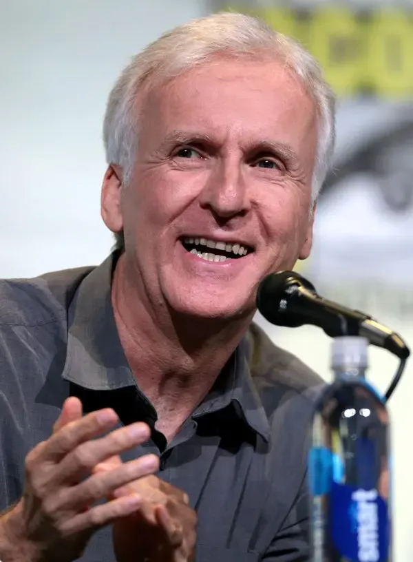 Kır saçlı yönetmen James Cameron, gri gömlekle mikrofon önünde gülümseyerek konuşurken