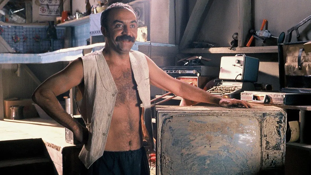 Vizontele filminden, eski radyolar ve aletlerle dolu bir atölyede duran bıyıklı, yelekli bir adam