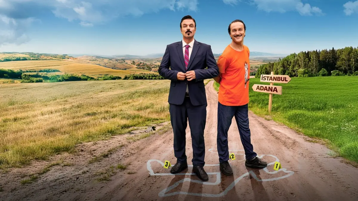 Geniş Aile 4: Cevahir & Ulvi filminden, kırsal bir toprak yolda tebeşirle çizilmiş olay yeri işaretlerinin üzerinde yan yana duran takım elbiseli Cevahir ve turuncu tişörtlü Ulvi