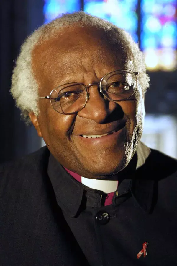 Gözlüklü ve beyaz saçlı Başpiskopos Desmond Tutu'nun koyu cüppesi ve rahip yakalığıyla vitray pencerenin önünde içtenlikle gülümsediği renkli fotoğrafı