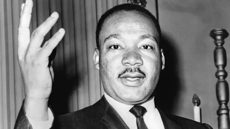 Martin Luther King Jr.'ın takım elbise ve kravatla sağ elini havaya kaldırıp hafifçe gülümseyerek kameraya baktığı siyah-beyaz fotoğrafı