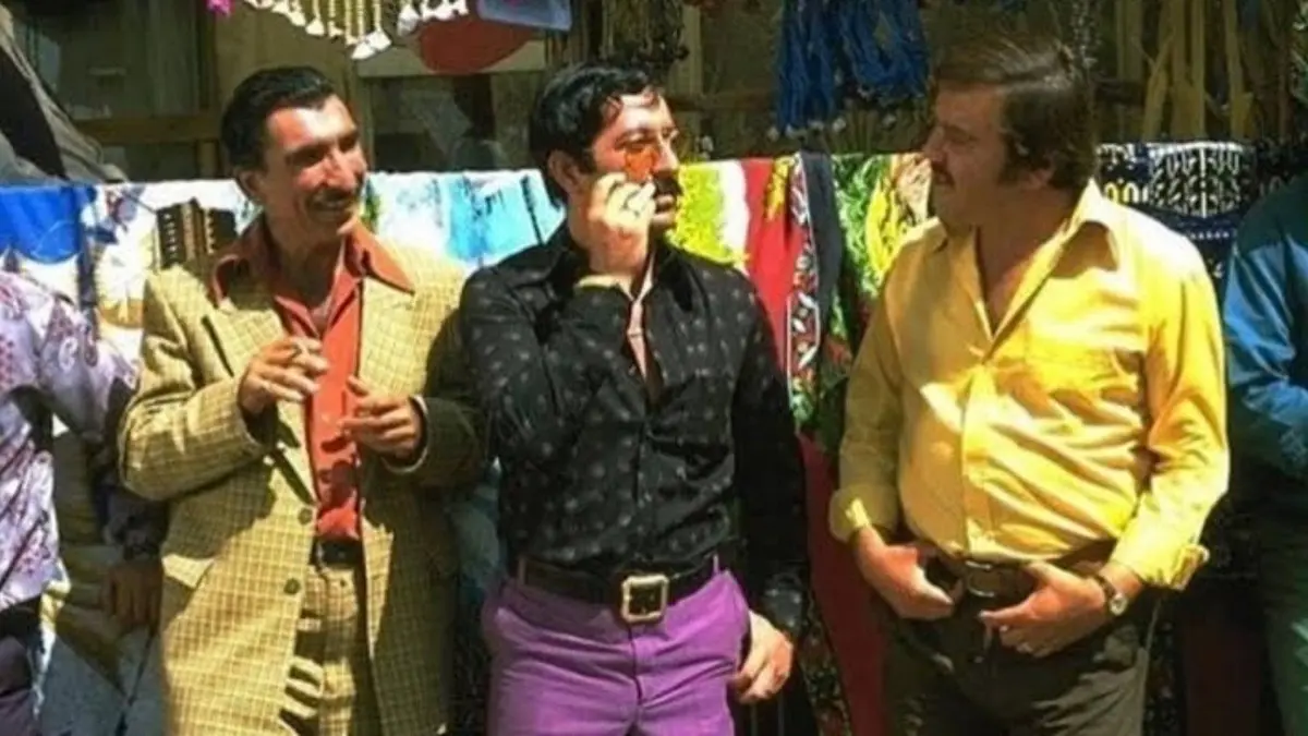 Vizontele filminden, kasaba çarşısında renkli kumaşların önünde duran ceketli, gömlekli üç adam
