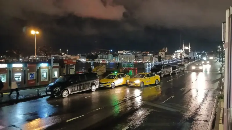 İstanbul’da yağmurlu bir gece: Galata Köprüsü çevresinde ıslak asfaltta sokak lambalarının ve araç farlarının parlak yansımaları, yolda taksiler ve otobüsler, arkada bulutlu gece göğü