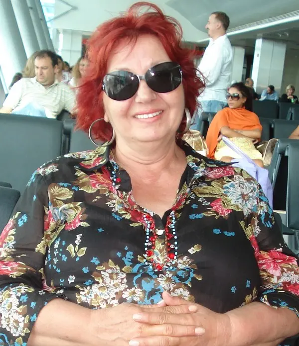 Kızıl saçlı, güneş gözlüklü, çiçek desenli koyu renk bluz giymiş oyuncu Güven Hokna'nın bir havalimanı bekleme salonunda gülümseyerek çekilmiş fotoğrafı
