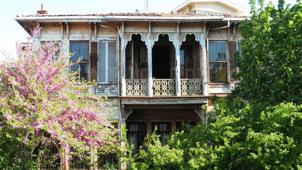 Beylerbeyi'nde, cumbalı ve ahşap oymalı balkonları olan eski iki katlı ahşap köşkün önden görünüşü; solda pembe çiçekli bir erguvan ağacı, önde yeşil ağaçlar