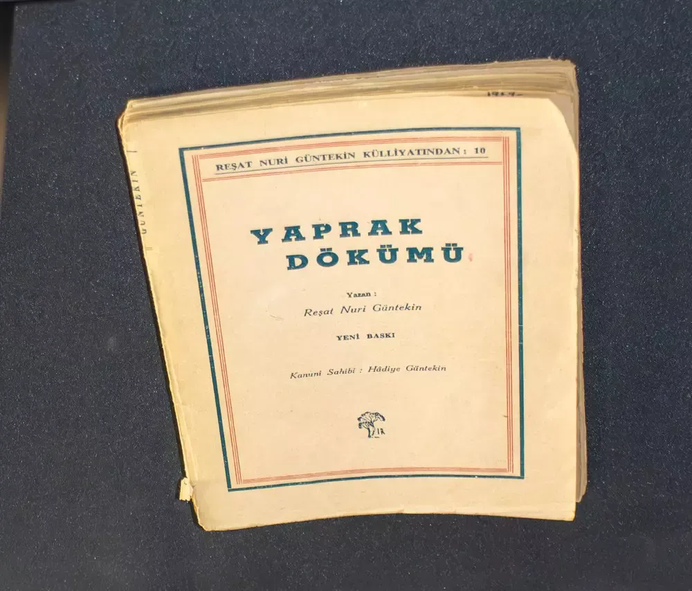 Koyu renk bir masa örtüsü üzerinde duran, sararmış kâğıt kapaklı eski bir Yaprak Dökümü baskısı; kapakta “Reşat Nuri Güntekin Külliyatından”, “Yazan: Reşat Nuri Güntekin” ve “Yeni Baskı” yazıları