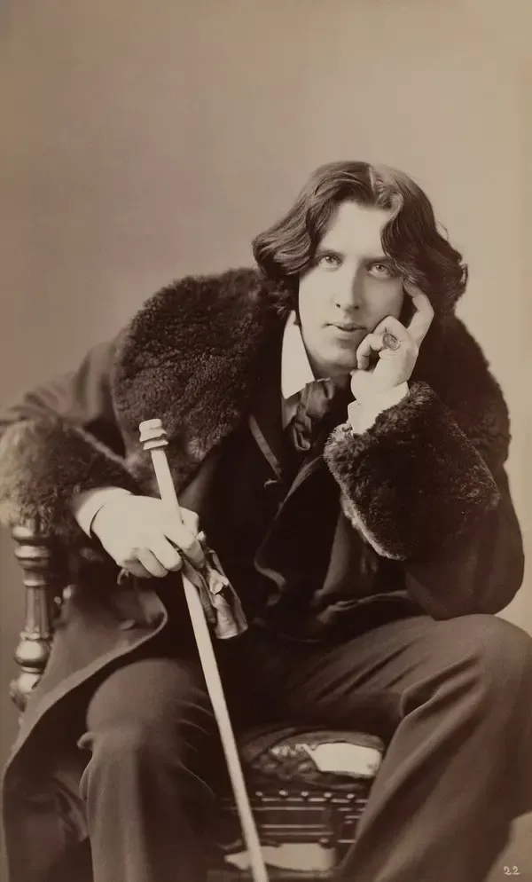 Uzun dalgalı saçlı genç Oscar Wilde'ın kürk yakalı paltosuyla bir koltukta oturup başını eline yasladığı, elinde bastonla kameraya baktığı sepya tonlu fotoğrafı