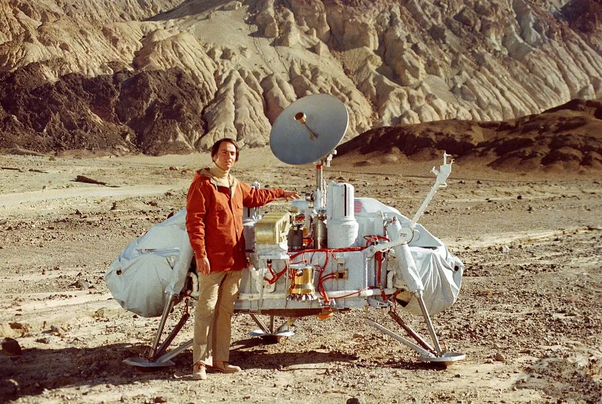 Kırmızı ceketli Carl Sagan'ın kayalık tepelerin önünde, çöl zemininde duran beyaz gövdeli ve çanak antenli Viking iniş aracının yanında ayakta durduğu renkli fotoğraf