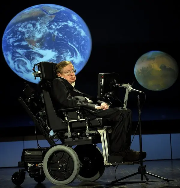 Tekerlekli sandalyesindeki Stephen Hawking'in karanlık bir sahnede, arkasındaki dev ekrana yansıtılan Dünya ve Mars görüntülerinin önünde konuşma yaptığı fotoğraf